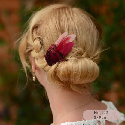 Blonde hair styled in an updo with a pink and burgundy feather accessory, blurred natural background