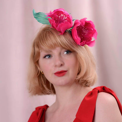 A woman with short blonde hair wearing a garden party headpiece of a side perching bright pink peony flower with green leaves made entirely with goose feathers, dressed in a red top against a soft pink background.