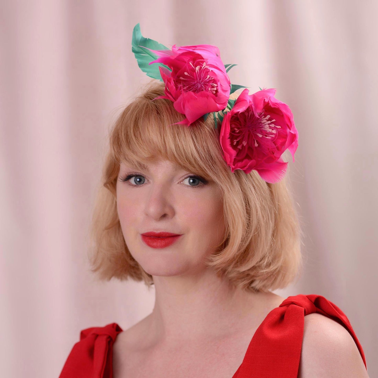 A woman with short blonde hair wearing a bright pink peony flower headpiece with green leaves made entirely with goose feathers, dressed in a red top against a soft pink background.