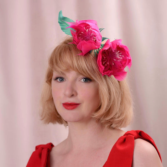 A woman with short blonde hair wearing a bright pink peony flower headpiece with green leaves made entirely with goose feathers, dressed in a red top against a soft pink background.