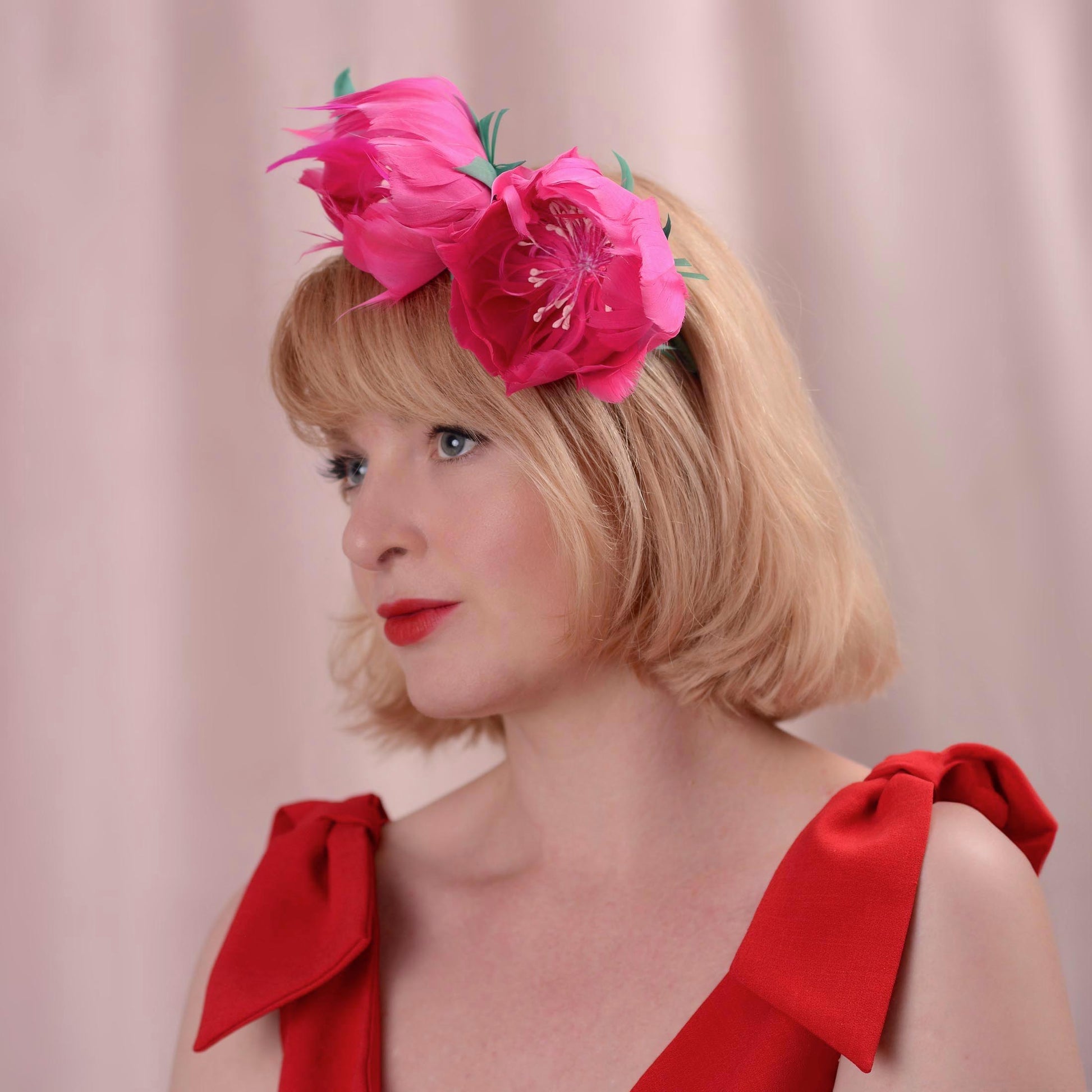 A blonde woman in a red dress with bow details wears a large pink floral headpiece with green leaves, posed against a soft pink backdrop.