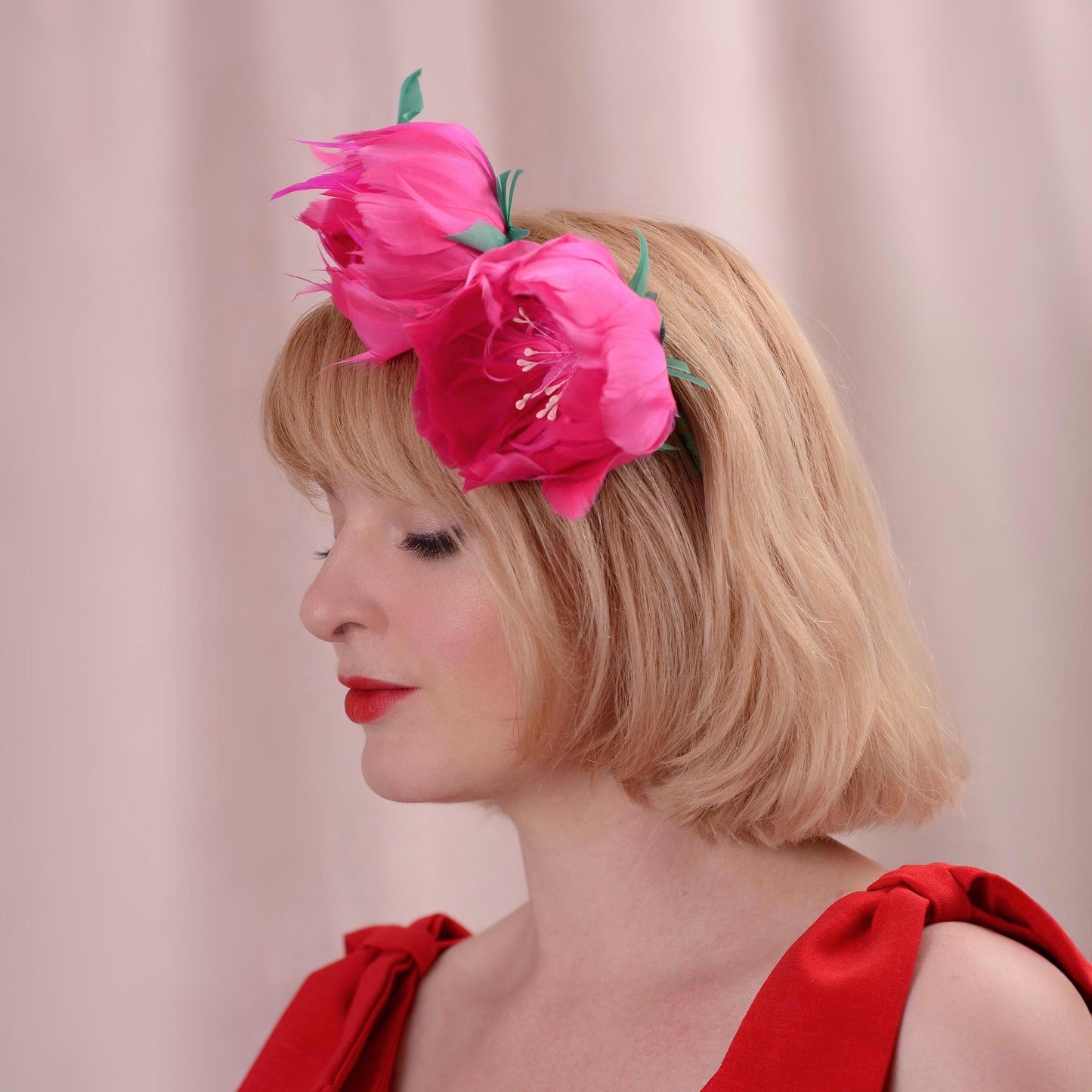 A blonde woman in profile wearing a bright pink floral hair accessory with green leaves made entirely of goose feathers, against a soft pink background.