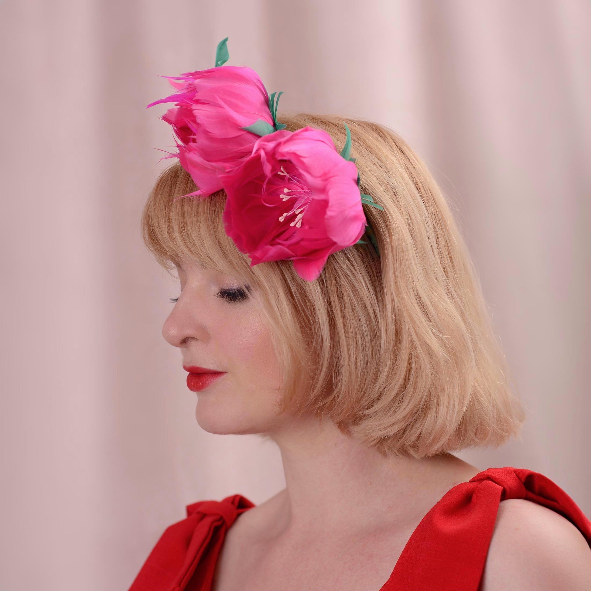 A blonde woman in profile wearing a bright pink floral hair accessory with green leaves made entirely of goose feathers, against a soft pink background.
