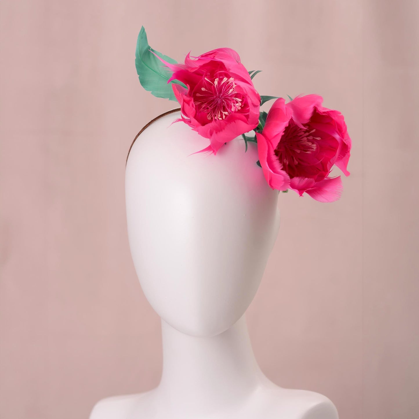 A white mannequin head and neck display wearing a thin headband adorned with two bright pink peony-style flowers and green leaves, set against a soft pink background.