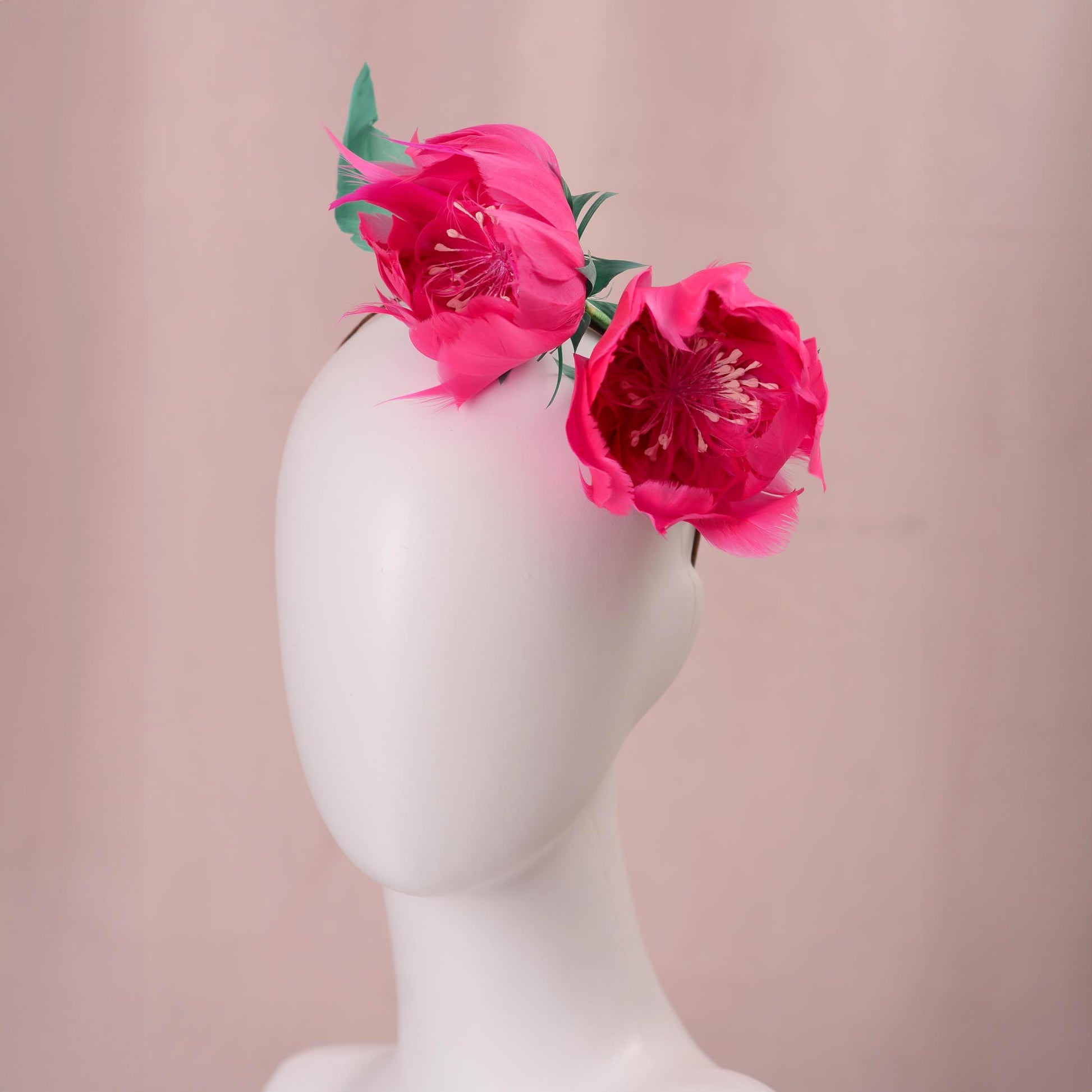 A  event headpiece consisting of pair of bright pink goose feather peony flowers with green leaves, displayed on a white mannequin head against a soft pink background.