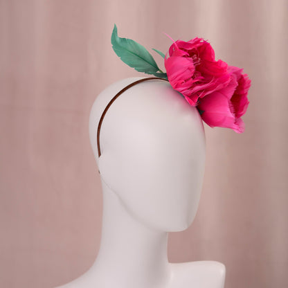A brown narrow headband with two large, vibrant magenta pink goose feather flowers and two green leaves, displayed on a white mannequin head against a soft pink backdrop.