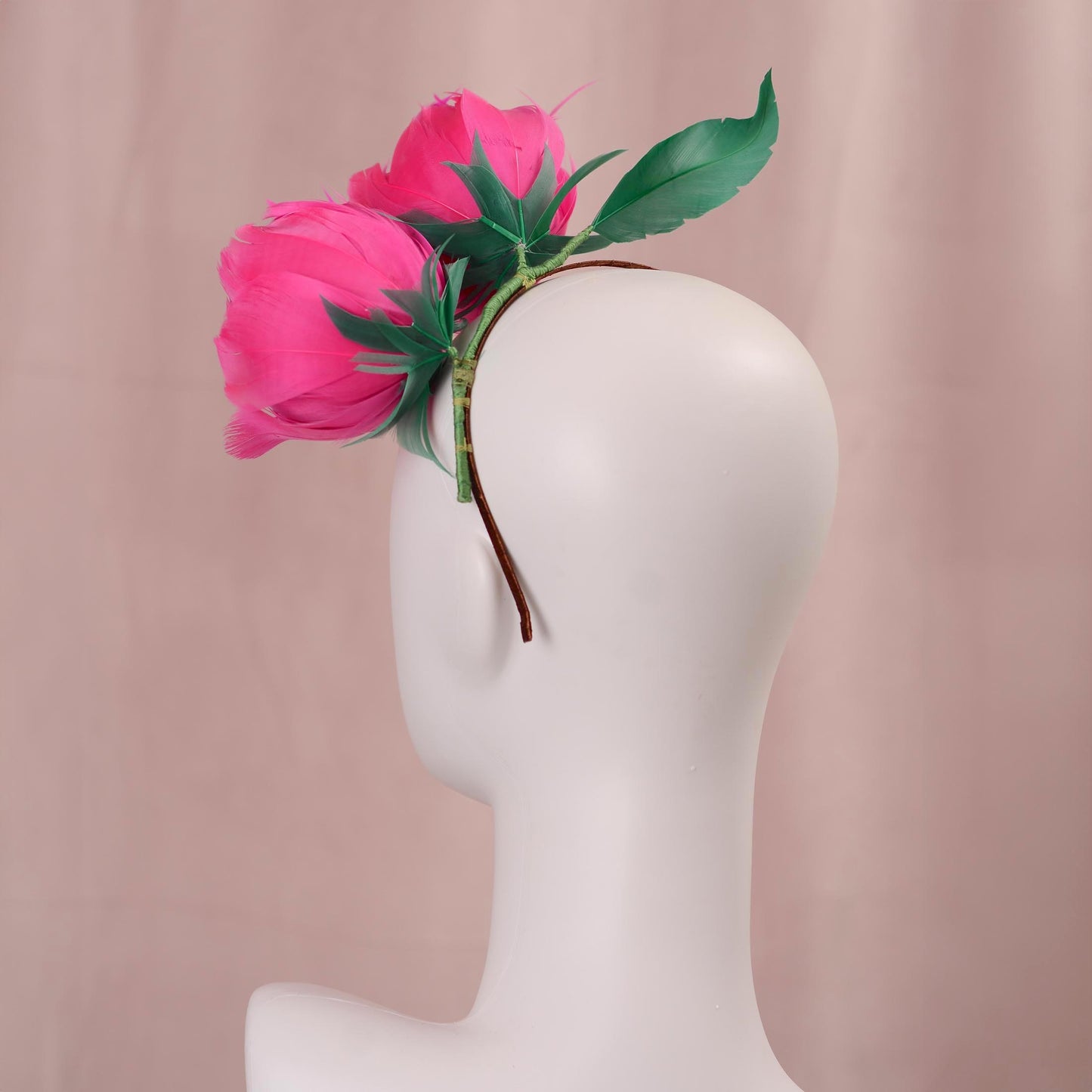 A wedding headpiece with vibrant pink goose feather peony flowers with green leaves styled on the side of a white mannequin head, set against a soft pink background. Back view showing couture detailing .