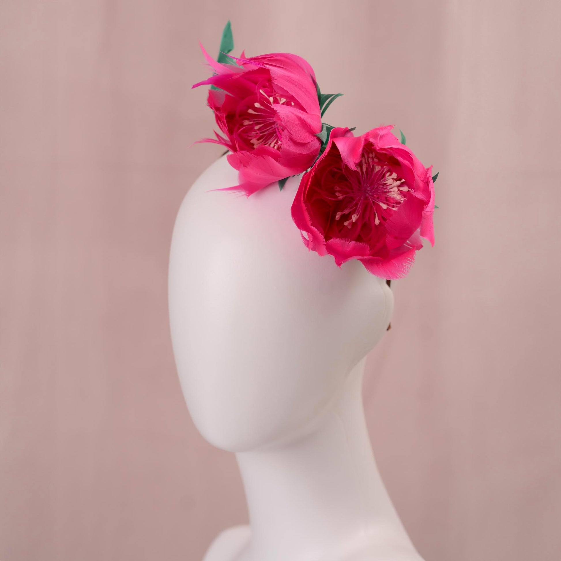 A wedding headpiece with vibrant pink goose feather peony flowers with green leaves styled on the side of a white mannequin head, set against a soft pink background.