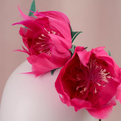 A wedding headpiece with vibrant pink goose feather peony flowers with green leaves styled on the side of a white mannequin head, set against a soft pink background. Close up view.
