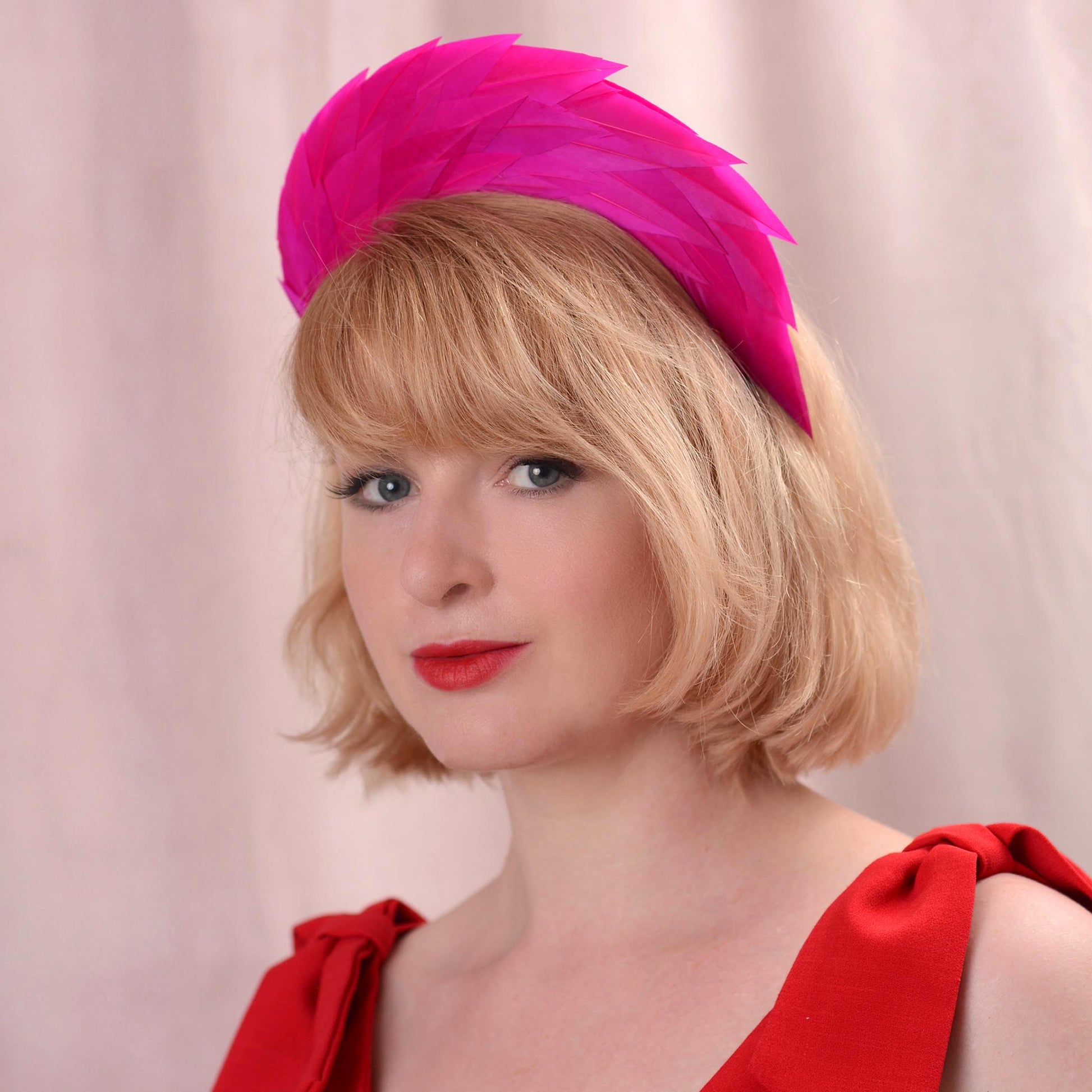 A woman with blonde hair wearing a bright pink feather fascinator headpiece and a red dress, photographed against a soft pink backdrop.