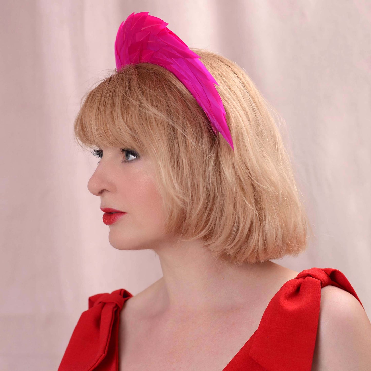 A woman with blonde hair wearing a bright pink feather fascinator headpiece and a red dress, photographed against a soft pink backdrop.