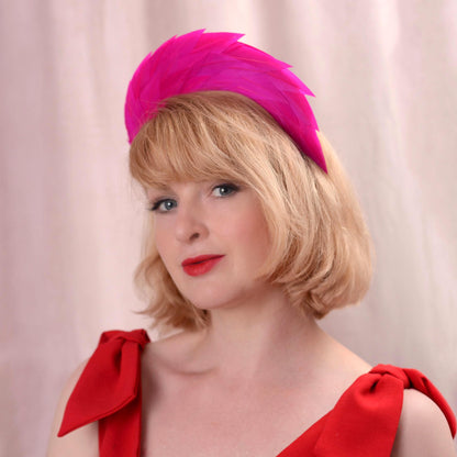 A woman with blonde hair wearing a bright pink feather fascinator headpiece and a red dress, photographed against a soft pink backdrop.