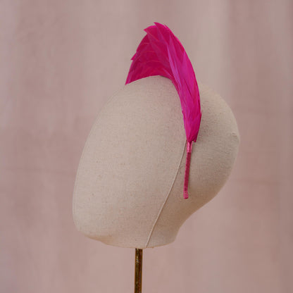  A bright hot pink headband featuring a tall, vertical arrangement of layered feathers that create a fan-like crest. The band itself is thin and wrapped in matching pink fabric, providing a secure base for the headpiece. Side view