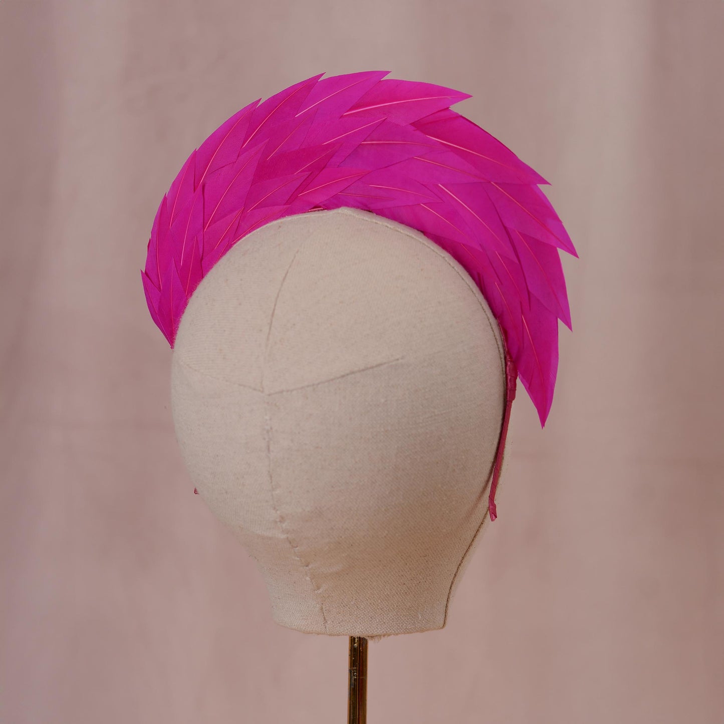 A bright pink feather headband or hair accessory displayed on a cream fabric mannequin head against a soft pink backdrop.