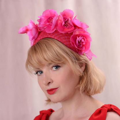 A woman with short blonde hair wears a bright pink and red fascinator headpiece with large fabric flowers and a red patterned band, against a soft pink background.