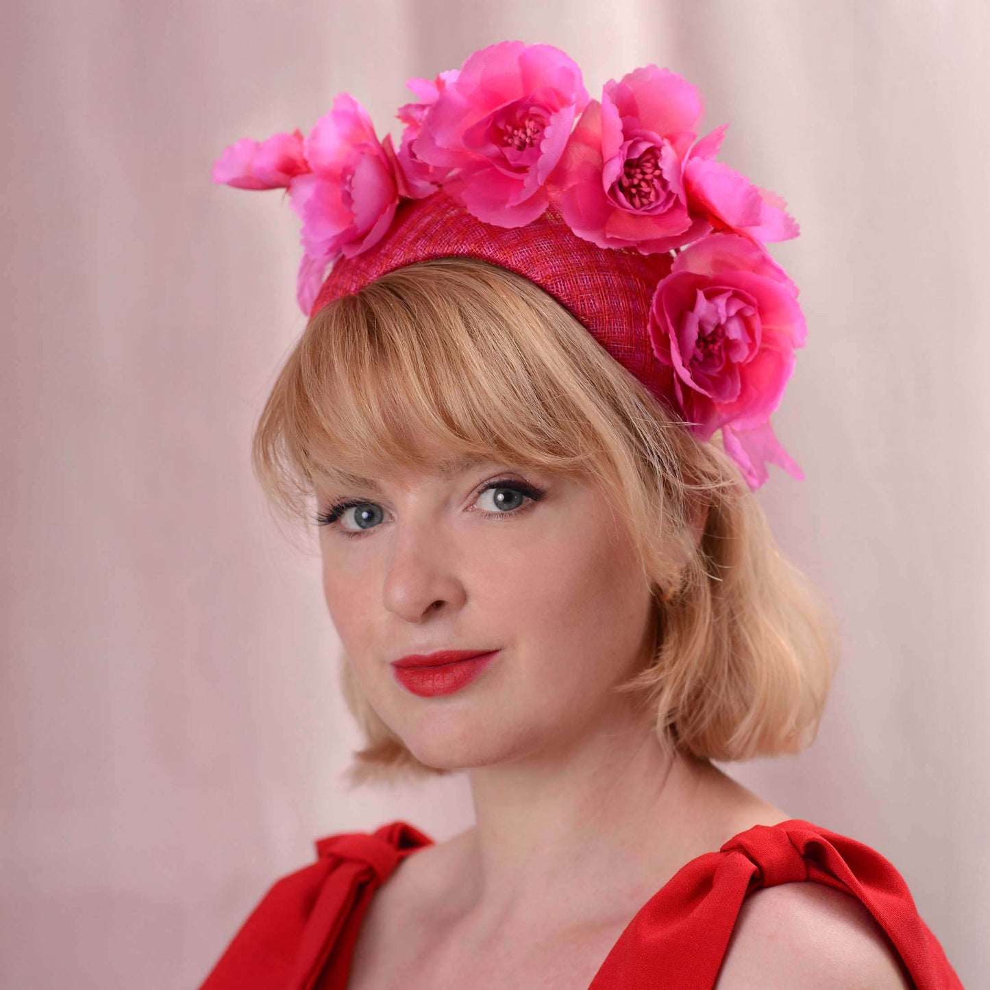 A woman with short blonde hair wearing a bright pink and red horse racing fascinator hat adorned with large artificial flowers. The hat is a 8cm wide bandeau headband style. She is posed against a soft pink studio backdrop.