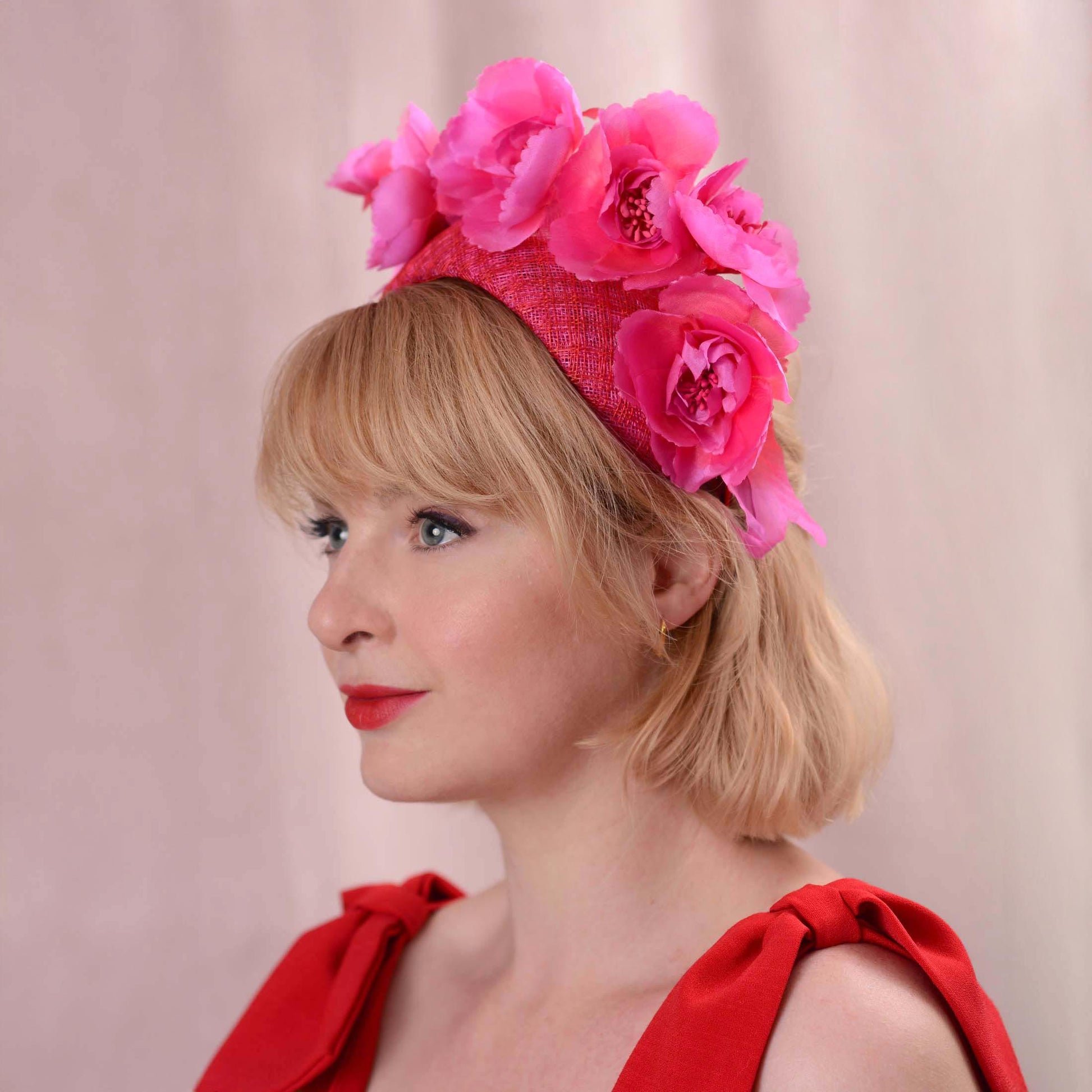 A woman in profile wearing a bright pink wedding fascinator headpiece with large fabric flowers and a pinka and red check patterned headband base, paired with a red sleeveless top  dress on a soft pink background.
