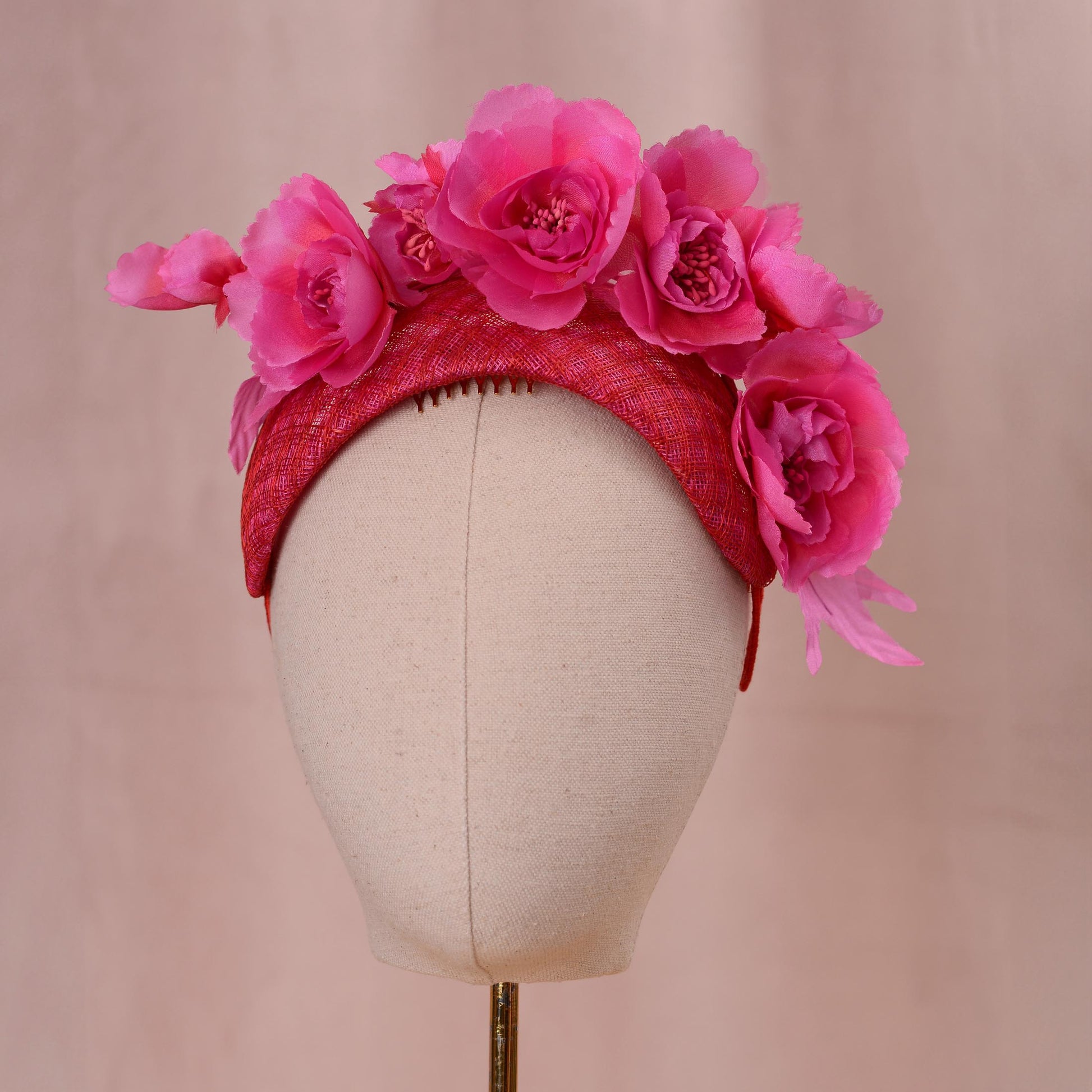 A bright pink and red fascinator headpiece in a bandeau headband style with large silk flowers mounted. Displayed on a beige fabric mannequin head against a soft pink background.
