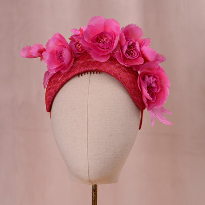 A bright pink and red fascinator headpiece in a bandeau headband style with large silk flowers mounted. Displayed on a beige fabric mannequin head against a soft pink background.