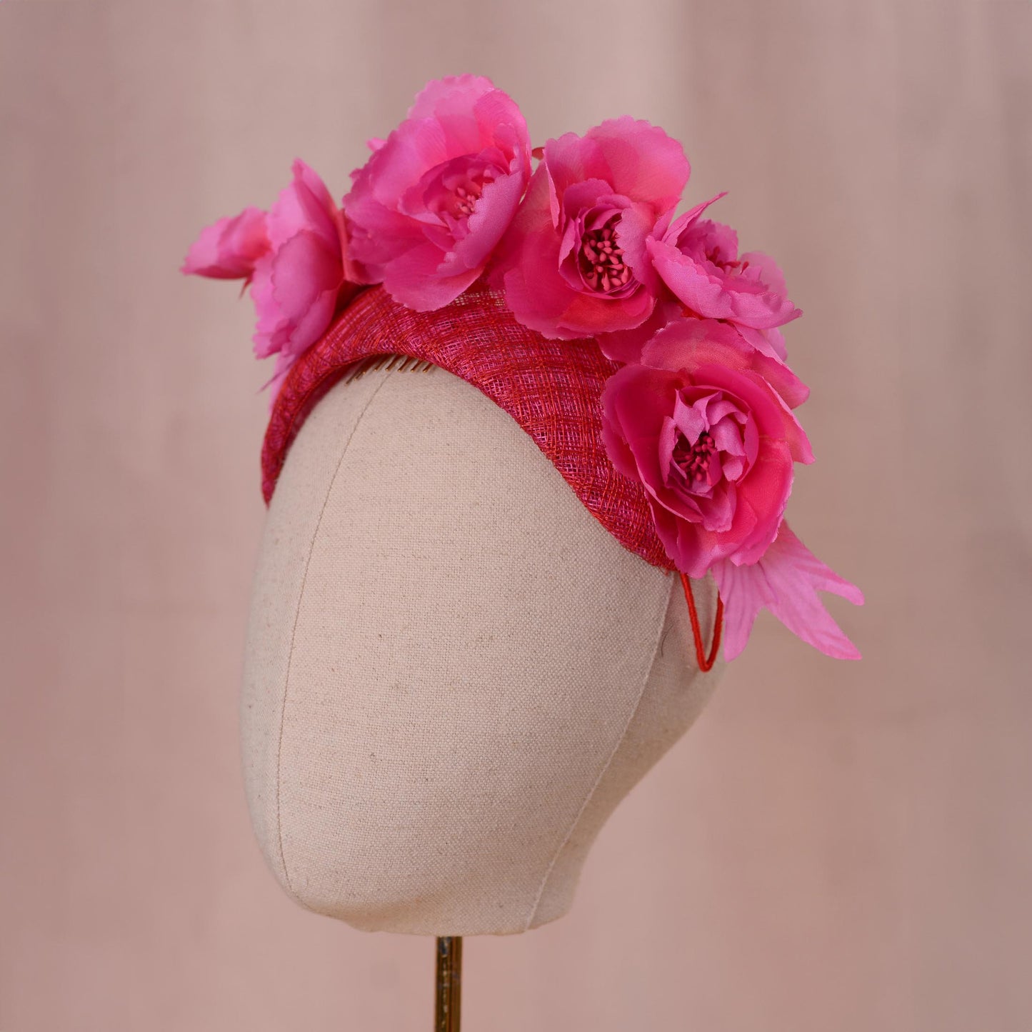 A bright pink and red Royal Ascot fascinator headpiece in a bandeau headband style with large silk flowers mounted. Displayed on a beige fabric mannequin head against a soft pink background.