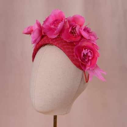 A bright pink and red Royal Ascot fascinator headpiece in a bandeau headband style with large silk flowers mounted. Displayed on a beige fabric mannequin head against a soft pink background.