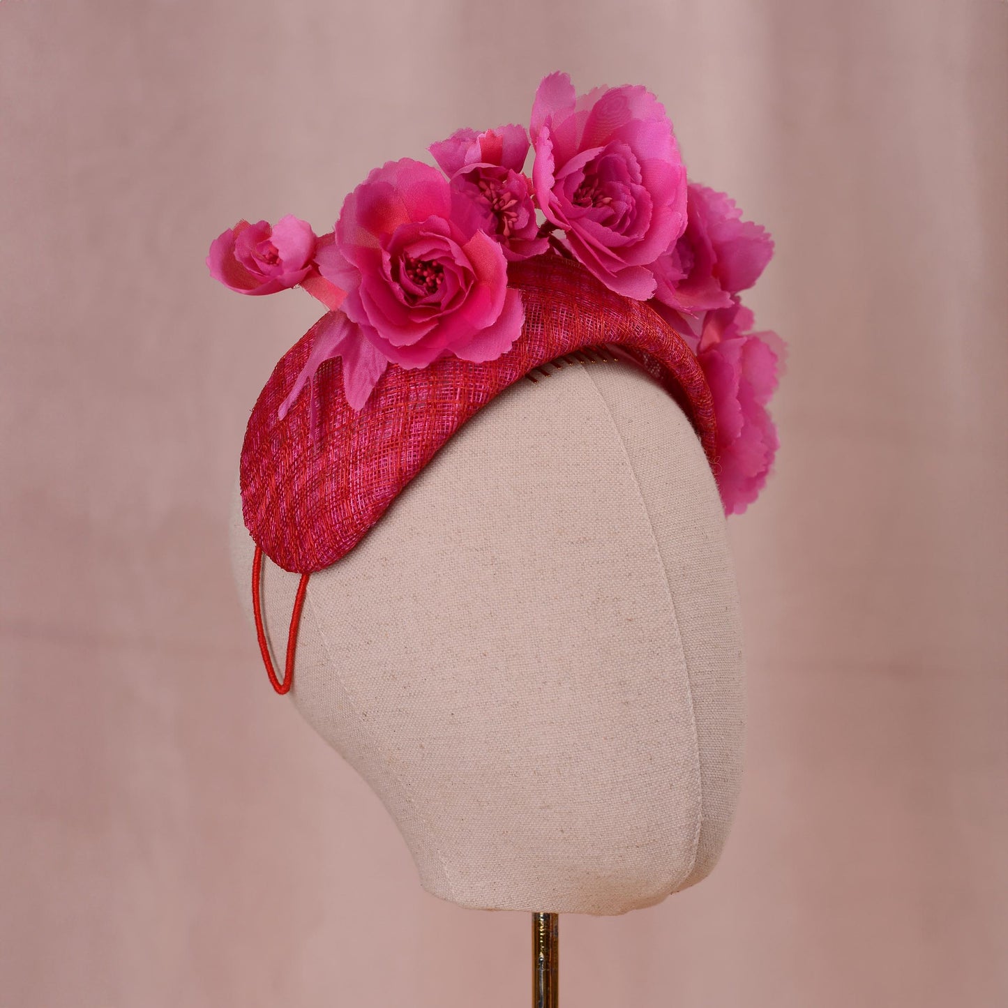 A bright pink and red fascinator headpiece in a bandeau headband style with large silk flowers mounted on a 8cm wide bandeau headband base in a pink and red checked straw. Displayed on a beige fabric mannequin head against a soft pink background.