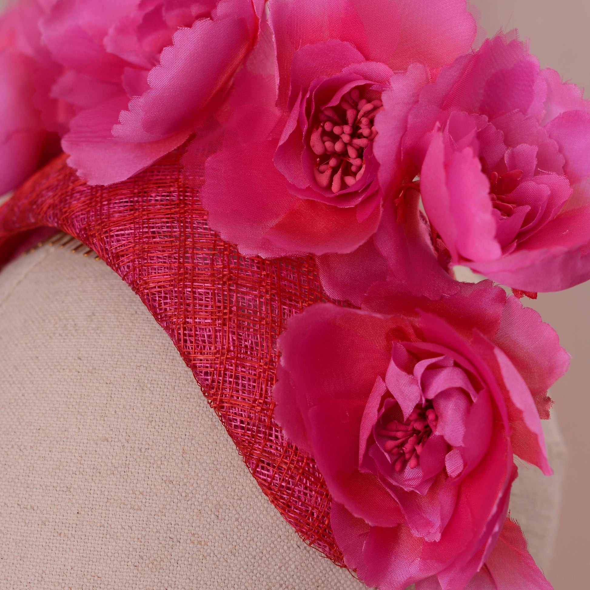 Close up of a bright pink and red fascinator headpiece in a bandeau headband style with large silk flowers mounted on a 8cm wide bandeau headband base in a pink and red checked straw. Displayed on a beige fabric mannequin head against a soft pink background.