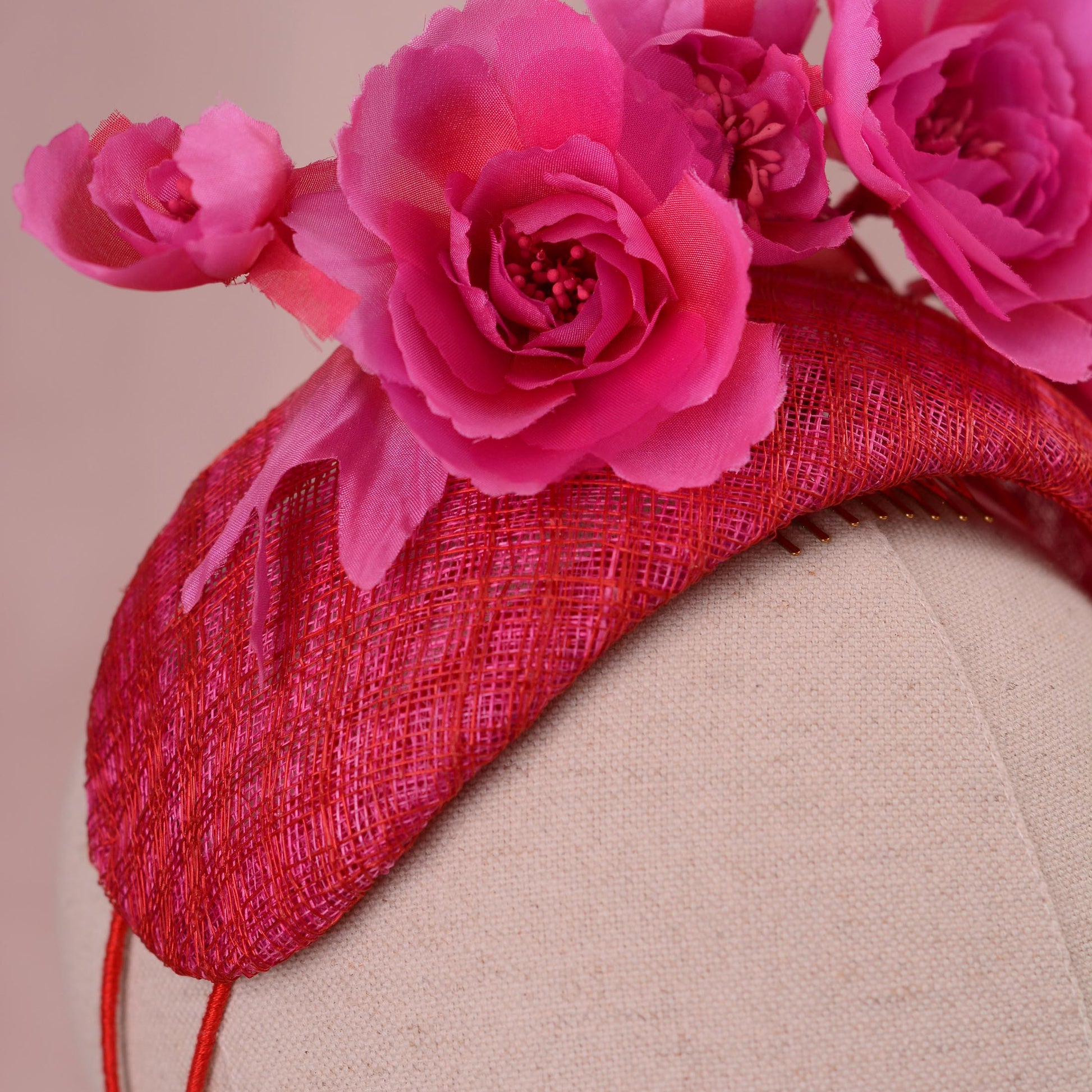 Close up of a bright pink and red fascinator headpiece in a bandeau headband style with large silk flowers mounted on a 8cm wide bandeau headband base in a pink and red checked straw. Displayed on a beige fabric mannequin head against a soft pink background.