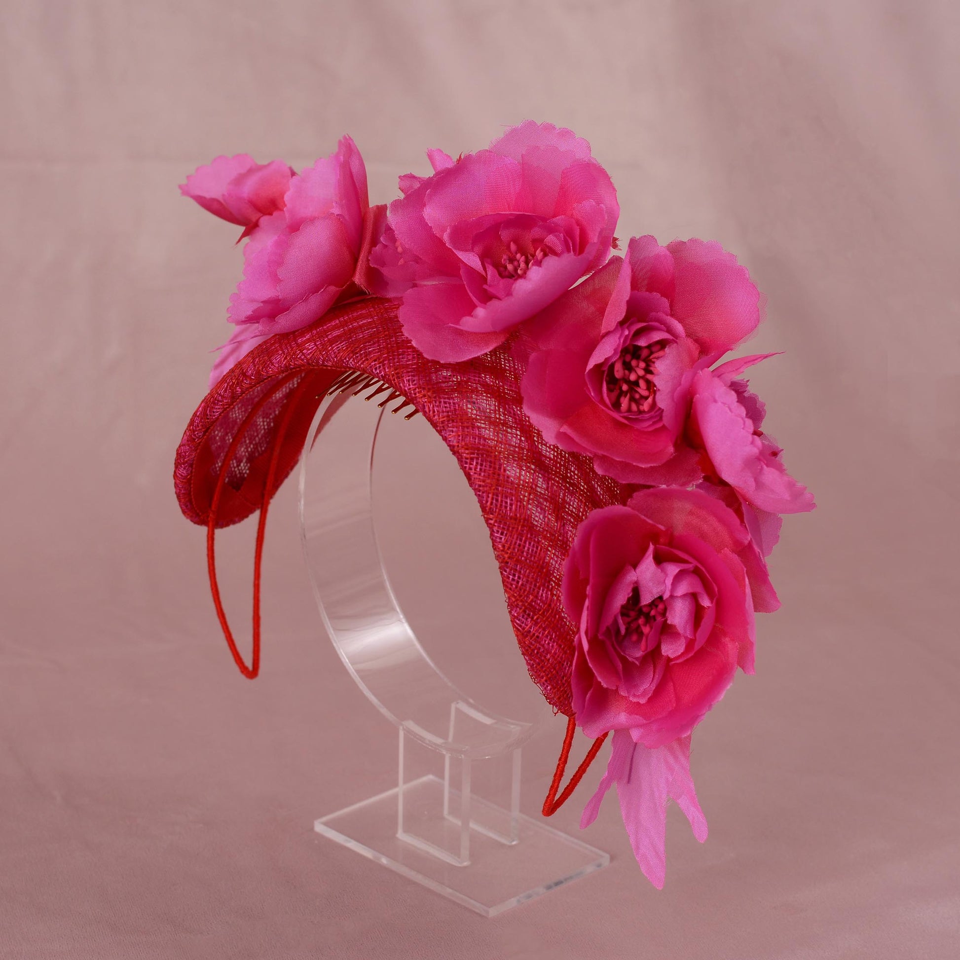 A pink and red wedding fascinator headpiece with large silk flowers on a pink and red straw bandeau headband base, displayed on a clear acrylic stand against a soft pink background.