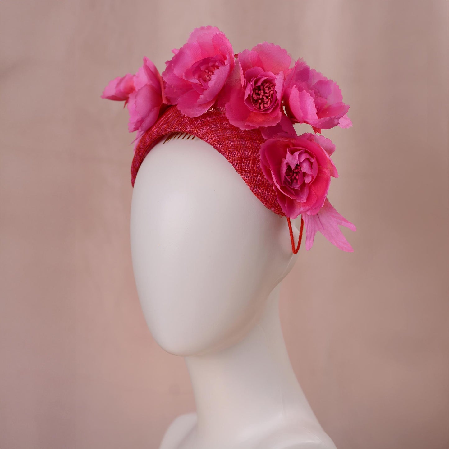 A bright pink and red silk floral fascinator headband displayed on a white mannequin head against a soft beige background.