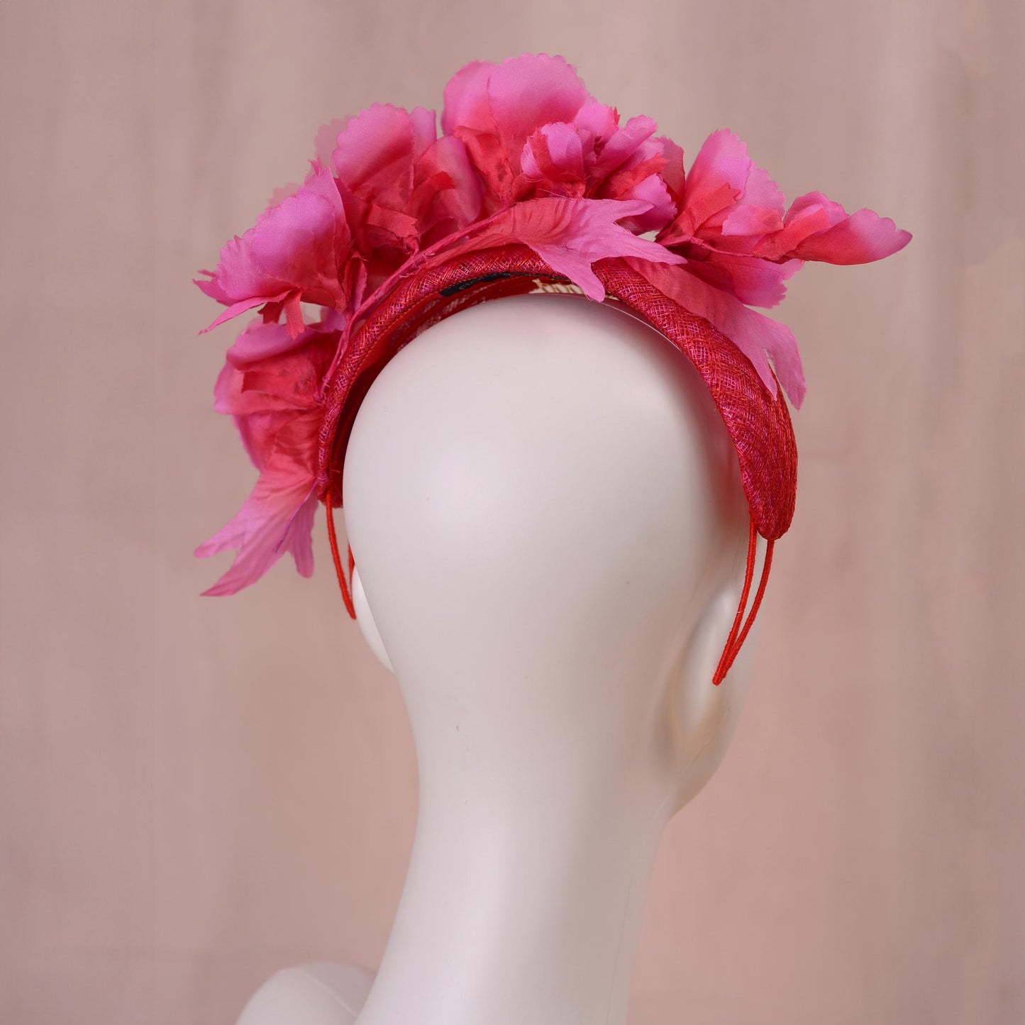 A bright pink and red silk floral fascinator headband displayed on a white mannequin head against a soft beige background.