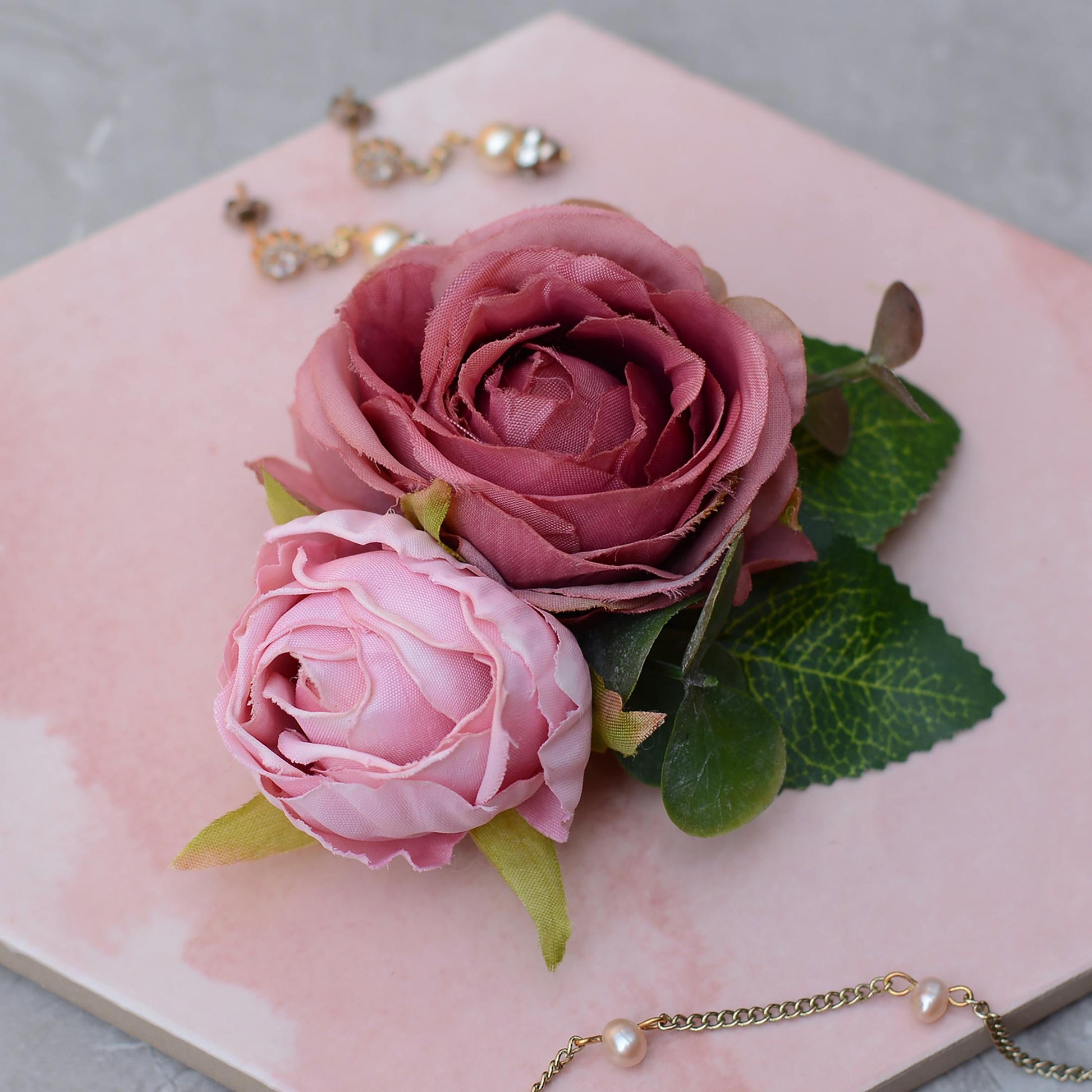 Bridal hair clip of realistic Pink roses with green leaves on a pink surface with jewelry in the background