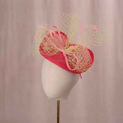  A vibrant tonal pink fascinator, presented on a mannequin head. The hat incorporates a vibrant yellow-green netted veil and is adorned with pink feathers. This headpiece is ideal for formal events and celebrations it is a suitable size for Royal Ascots Royal Enclosure rules