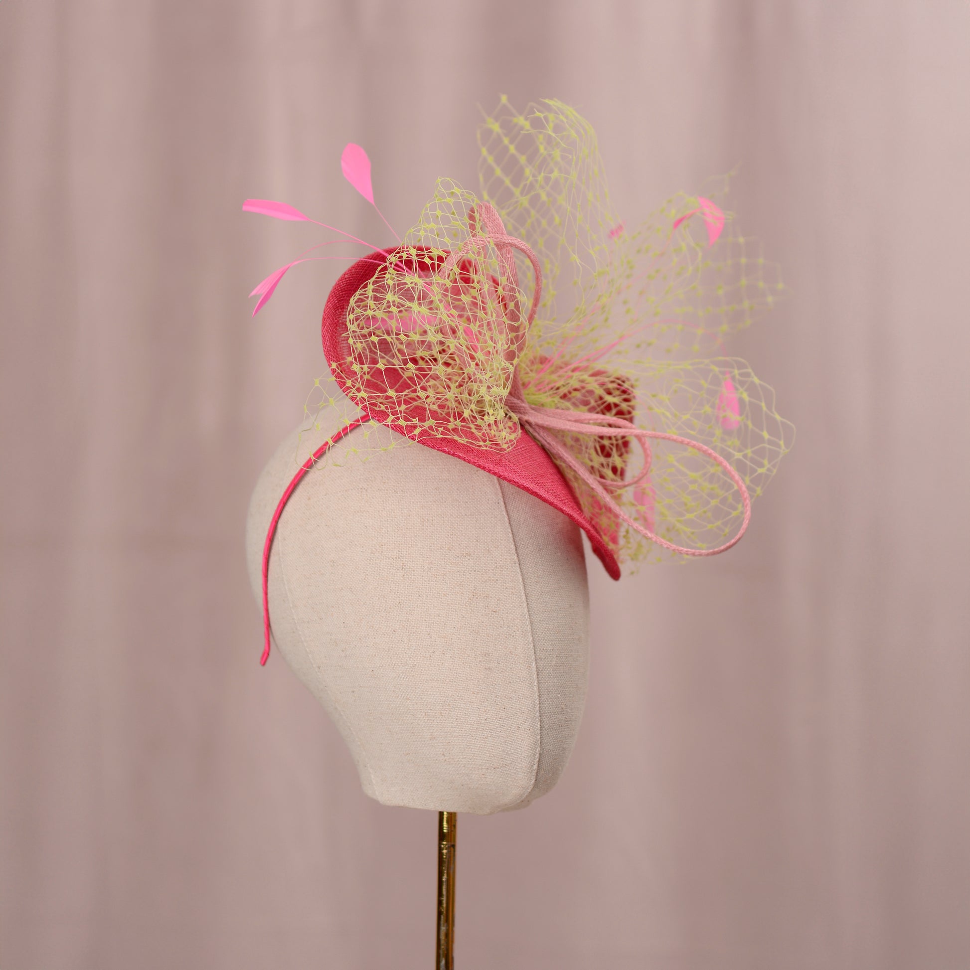 A summery pink headpiece with a decorative design. The headpiece features a pink headband, a curved pink base, and a layered design with a yellow net overlay. Pink feathers and accents complete the look. The headpiece is displayed on a mannequin head. Ideal for weddings and horse racing events sucha as the kentucky derby