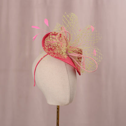 A summery pink headpiece with a decorative design. The headpiece features a pink headband, a curved pink base, and a layered design with a yellow net overlay. Pink feathers and accents complete the look. The headpiece is displayed on a mannequin head. Ideal for weddings and horse racing events sucha as the kentucky derby