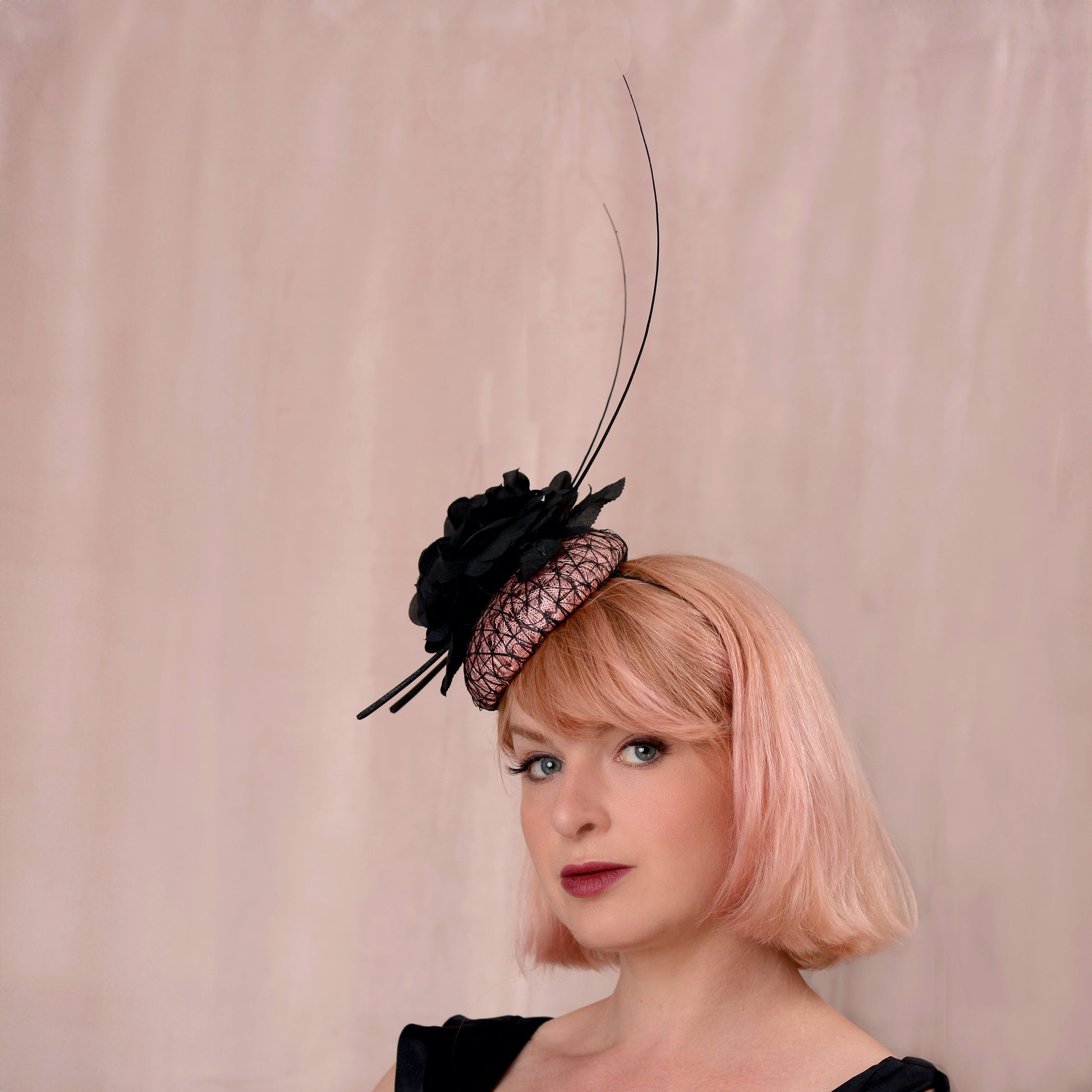 A woman with a pink bob haircut wearing a black and blush pink fascinator hat with a tall feather accent, photographed against a soft beige backdrop.