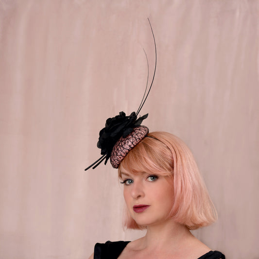 A woman with a pink bob haircut wearing a black and blush pink fascinator hat with a tall feather accent, photographed against a soft beige backdrop.