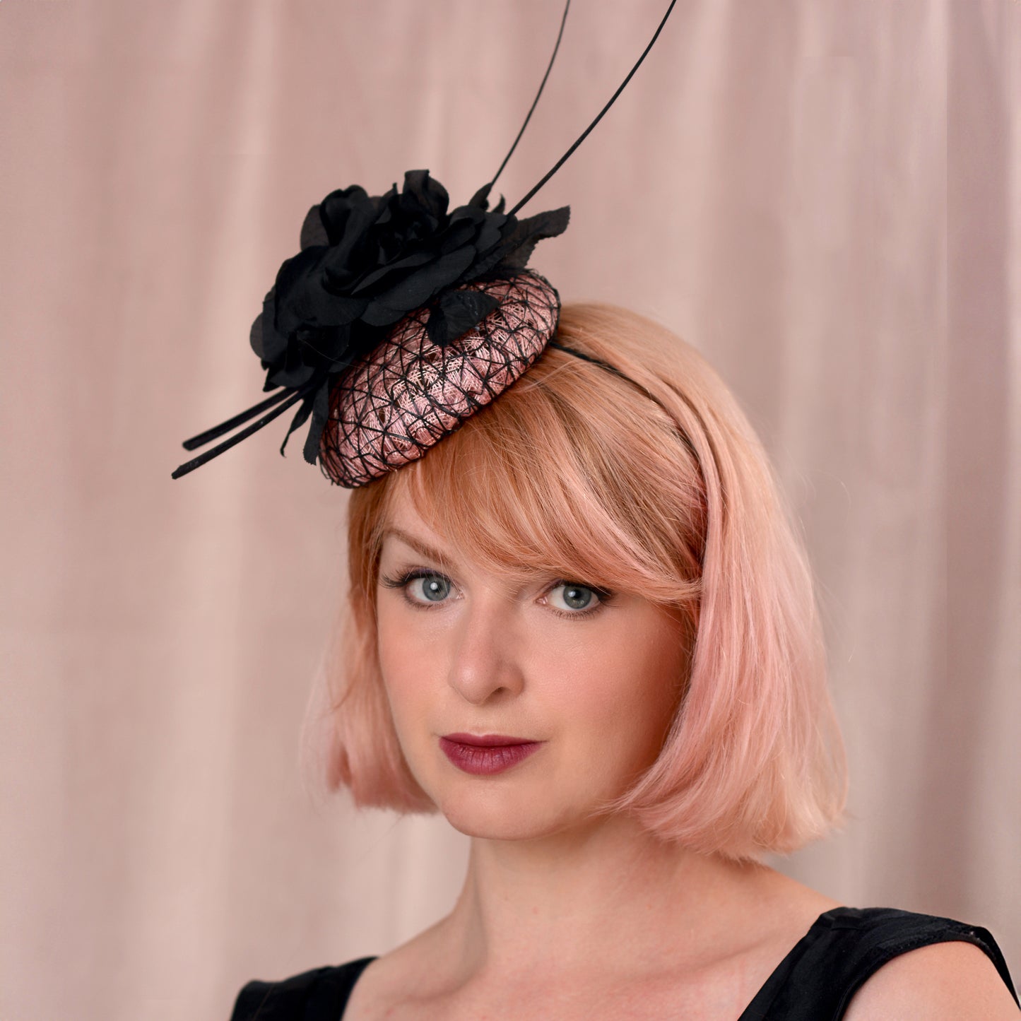 A woman with a short pink-blonde bob wears a black fascinator hat with black flowers and black mesh detailing over a blush pink button base, posed against a soft beige curtain backdrop.