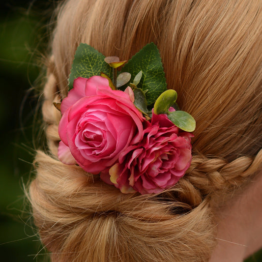 Blonde hair styled in an updo decorated with a hair clip of realistic pink fabric flowers and green leaves against a blurred natural background