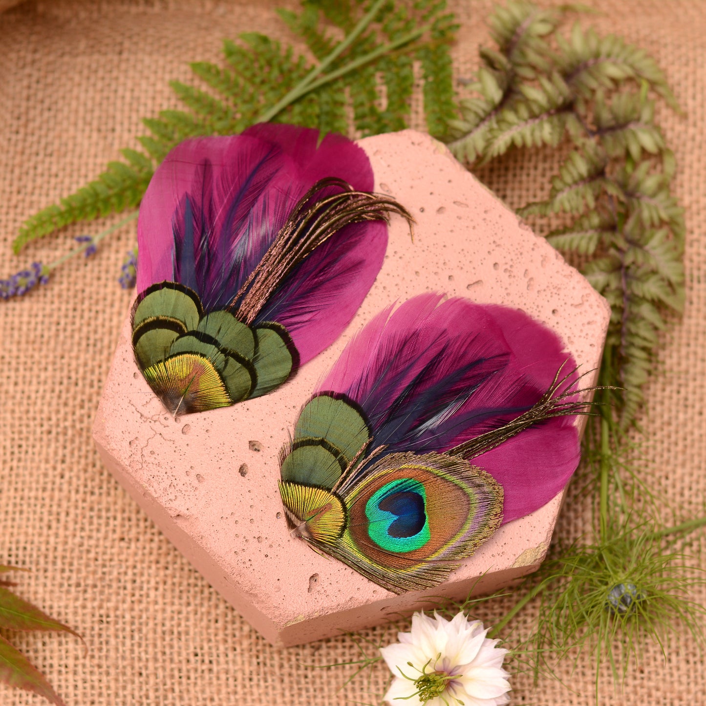 pair of matching purple and peacock feather hair clips on a pink stone and textured fabric surrounded by flowers