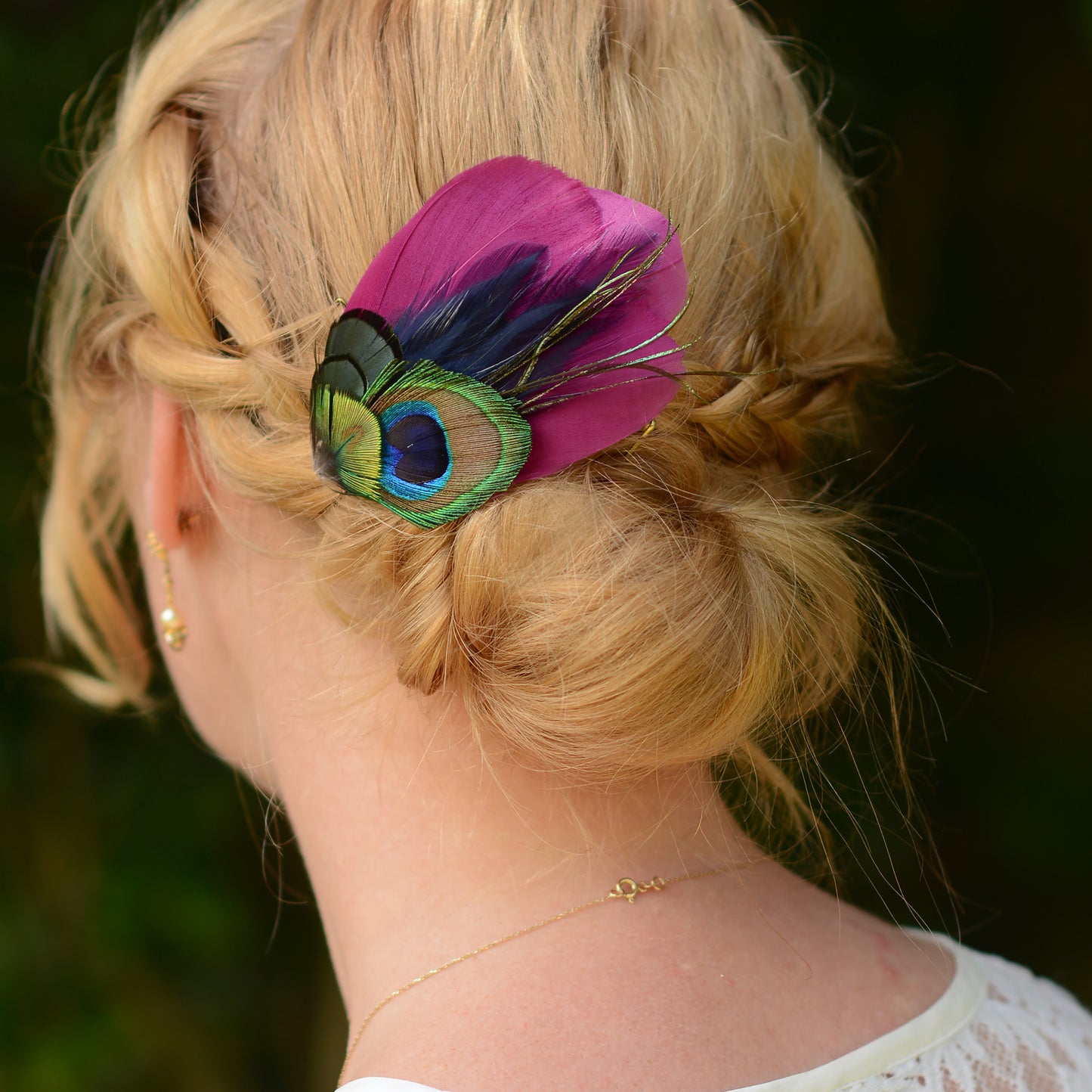 hair clip accessory with purple and green peacock eye feathers on the back or a woman's head with a braided low bun up-do
