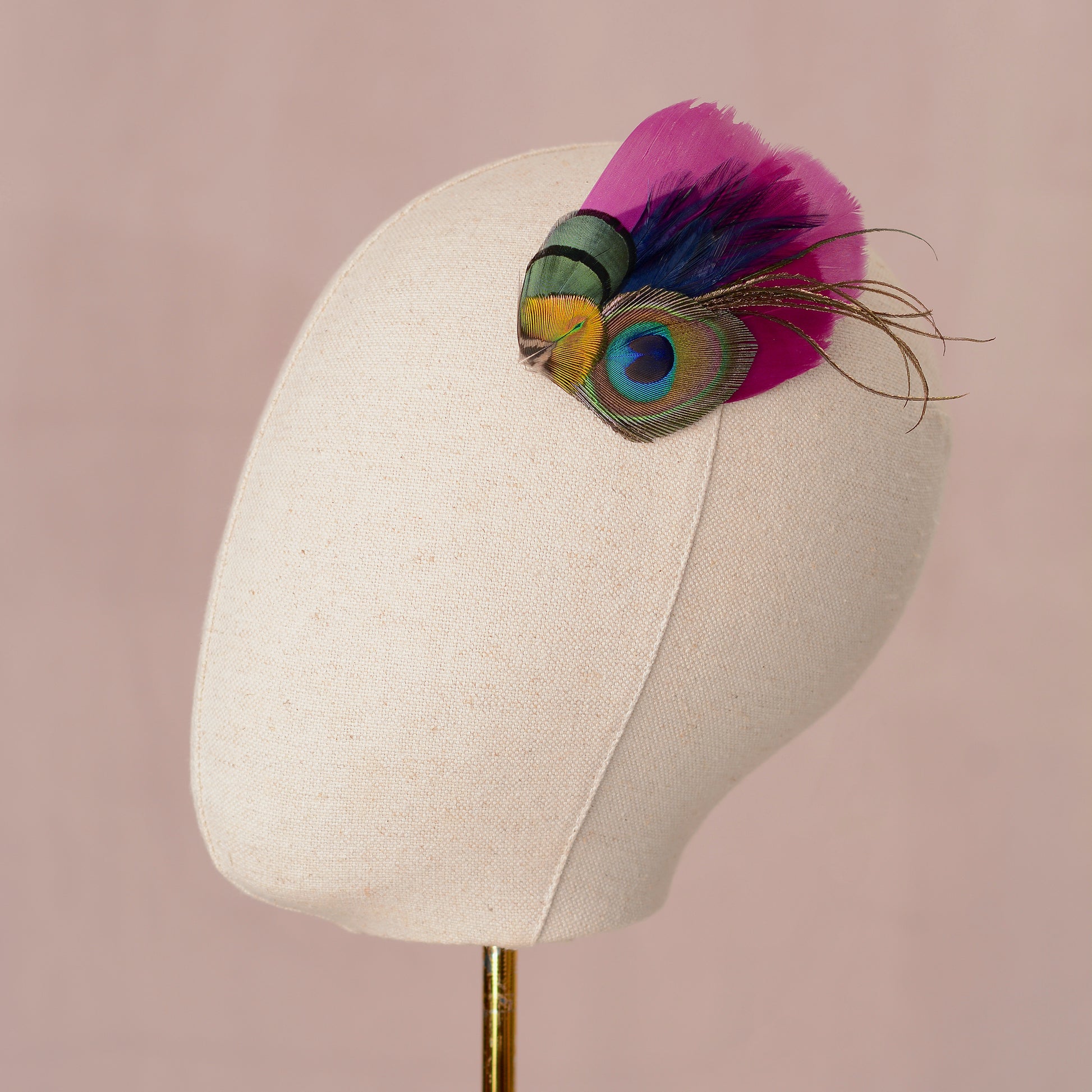 Mannequin head with a decorative peacock feather hair clip placed on the side of the head as a fascinator on a pink background