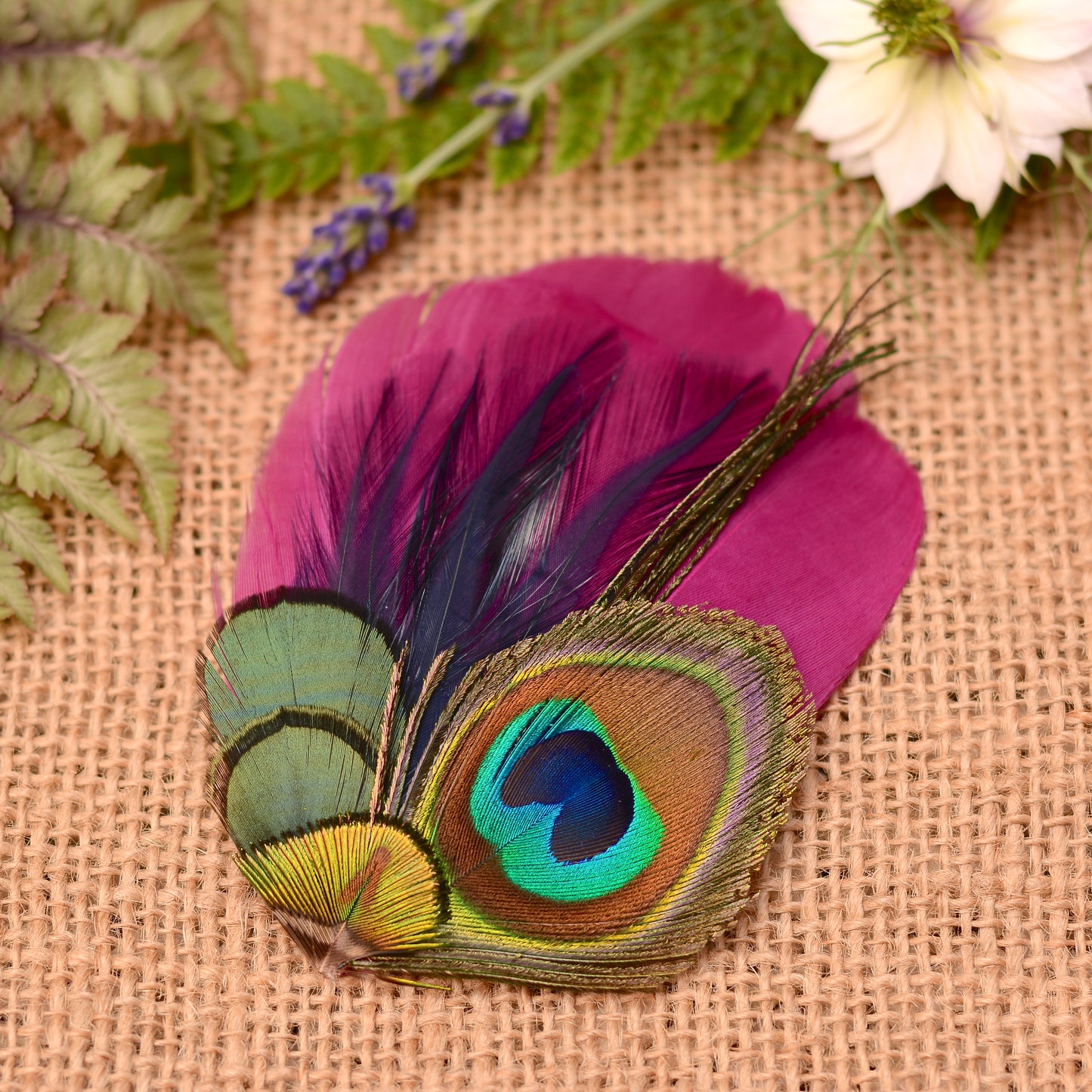 Purple  and peacock eye feather hair clip on a textured fabric background with flowers and plants.