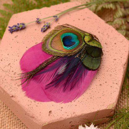Purple and peacock eye feather hair clip on a pink textured stone background with flowers and plants.