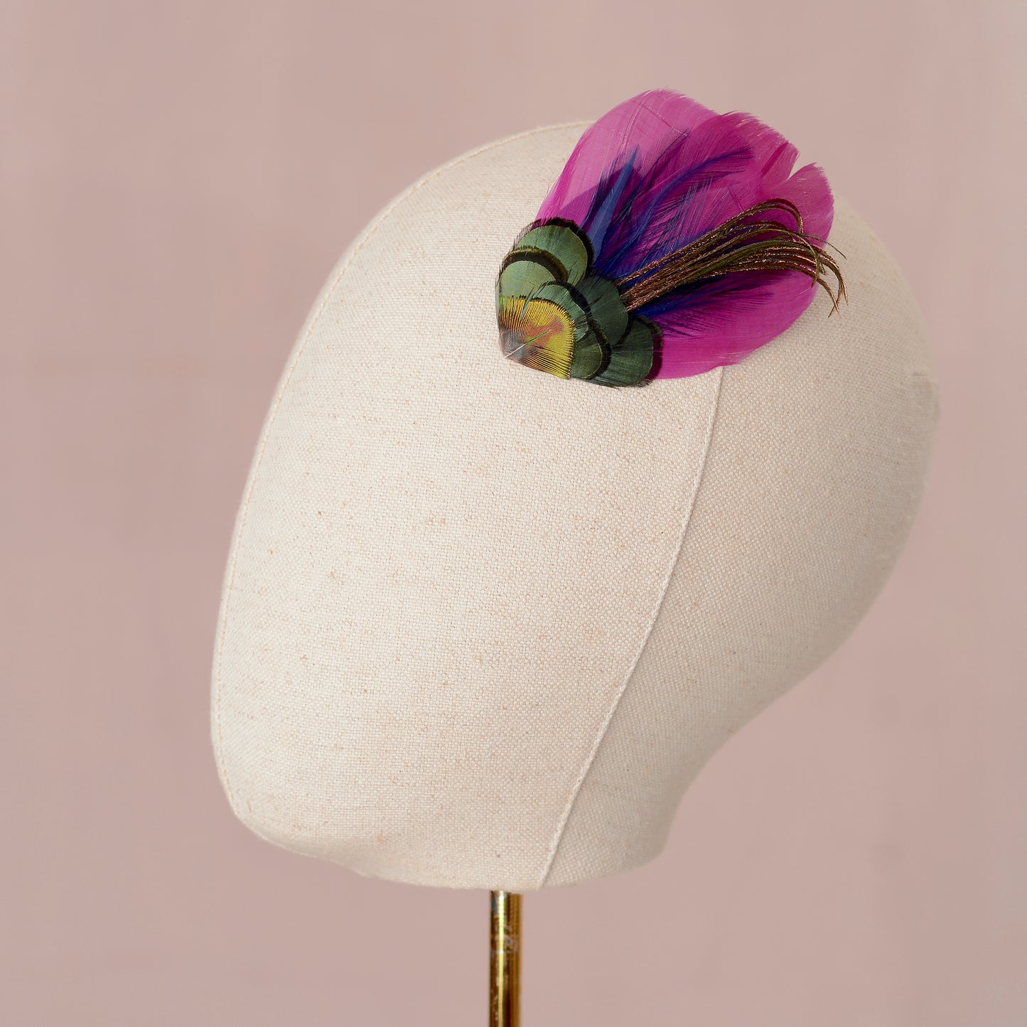 Mannequin head with a decorative peacock feather hair clip placed on the side of the head as a fascinator on a pink background