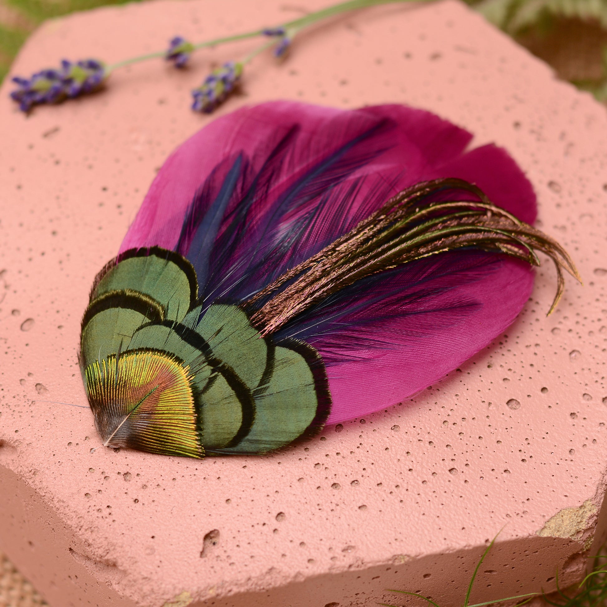 Close-up purple feather hair clip on a pink textured stone background with flowers and plants.