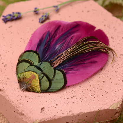 Close-up purple feather hair clip on a pink textured stone background with flowers and plants.