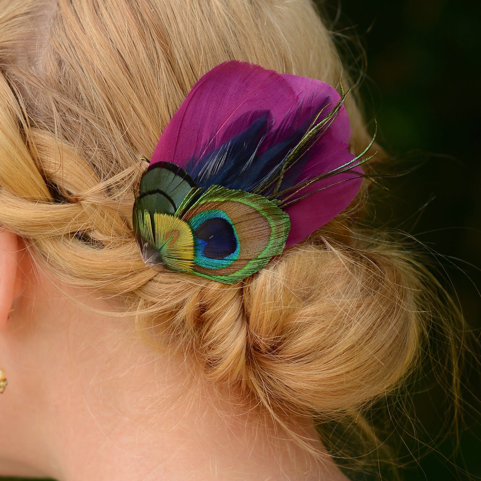 close up of hair clip accessory with purple and green peacock eye feathers on the back or a woman's head with a braided low bun up-do