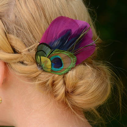 close up of hair clip accessory with purple and green peacock eye feathers on the back or a woman's head with a braided low bun up-do