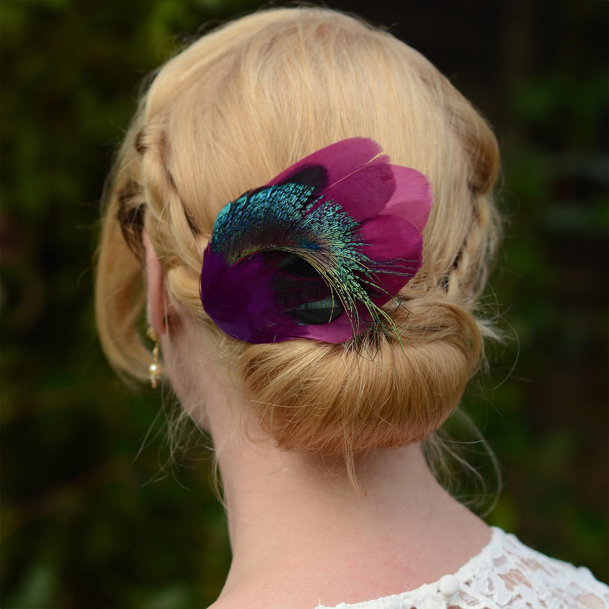 A blonde woman shown from the back wearing a decorative feather hair accessory in deep purple and iridescent teal peacock feather, set against a blurred green outdoor background.